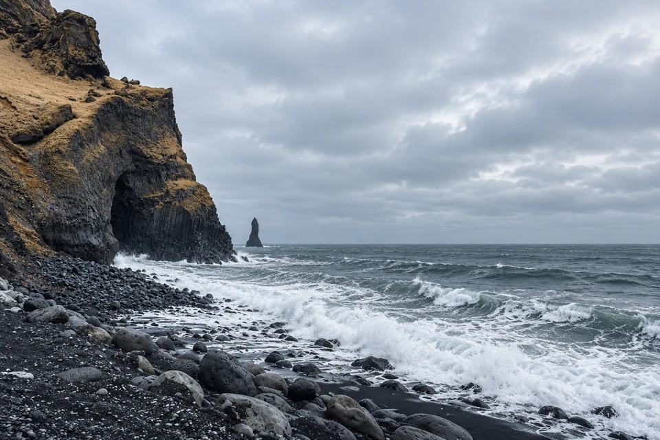 Reynisfjara's dark columnar basalt cliff rises on the left above black pebbles and rounded boulders, with white surf and an offshore rock stack beyond.
