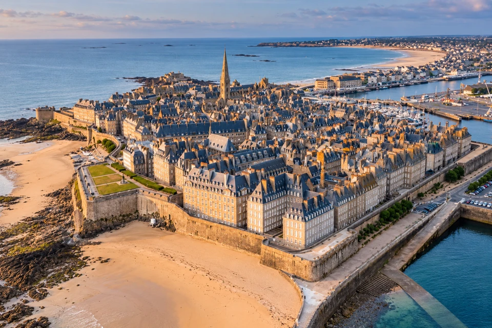 An elevated coastal view shows Saint-Malo's stone ramparts, dense slate-roofed old town, cathedral spire and surrounding sand and sea.