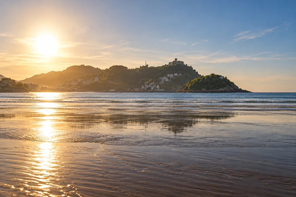 Wet sand reflects low sunlight on San Sebastian's La Concha beach, with Santa Clara island and the hillside behind the bay visible across the water.