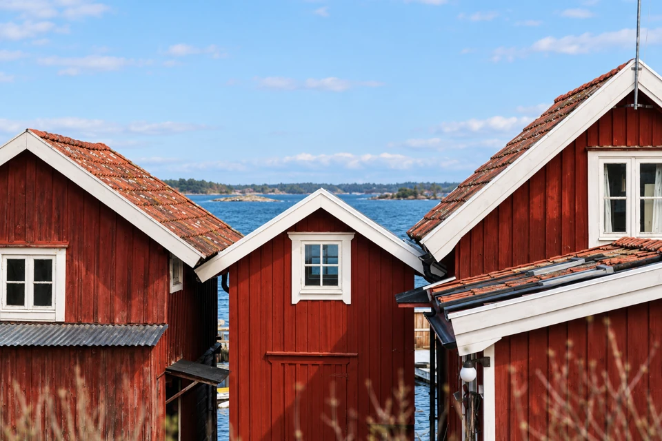 Three red timber waterfront buildings with white trim in Sandhamn, with Baltic water and low islands beyond.