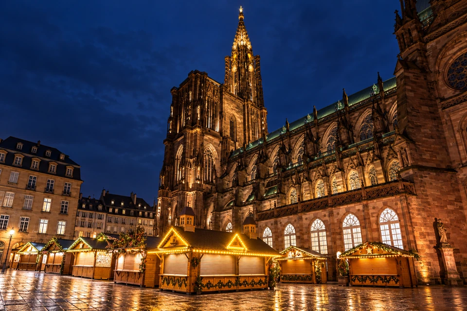 Strasbourg Cathedral illuminated above wooden Christmas-market chalets on a winter evening.