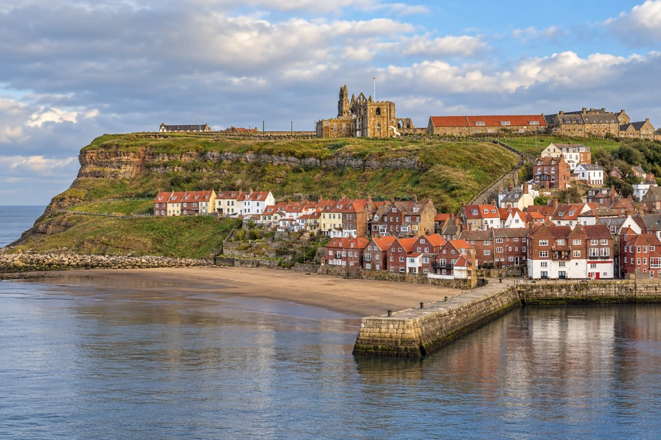 Whitby's lower harbor and red-roofed waterfront houses beneath the grassy East Cliff and historic stone skyline.